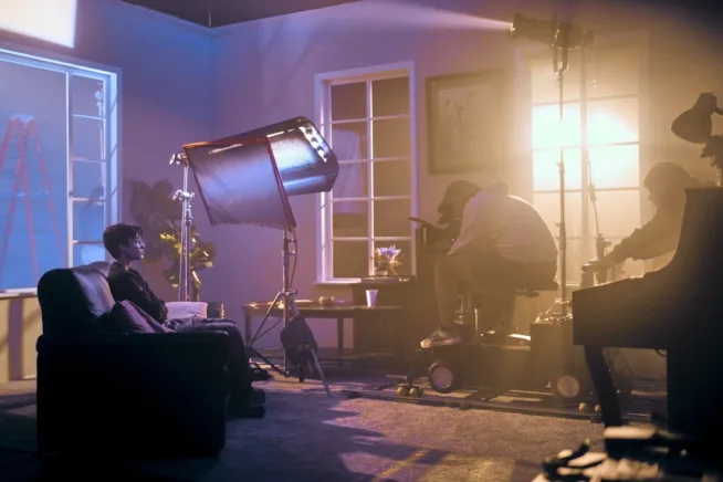 Dimly lit film set with a person sitting on a sofa, surrounded by cameras and lighting equipment. Crew members adjust gear, creating a focused atmosphere.